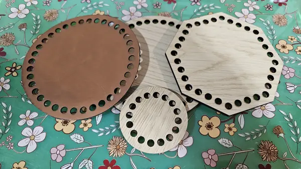 Three laser-cut crochet bases on a green floral fabric: a round Buckskin black glossy leatherette base, an octagonal TruFlat oak plywood base, and a smaller round TruFlat oak plywood base, each with a perimeter ring of crochet holes