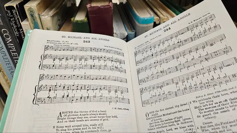 Un himnario abierto con notación musical grabada y versos, con libros de música en el estante detrás